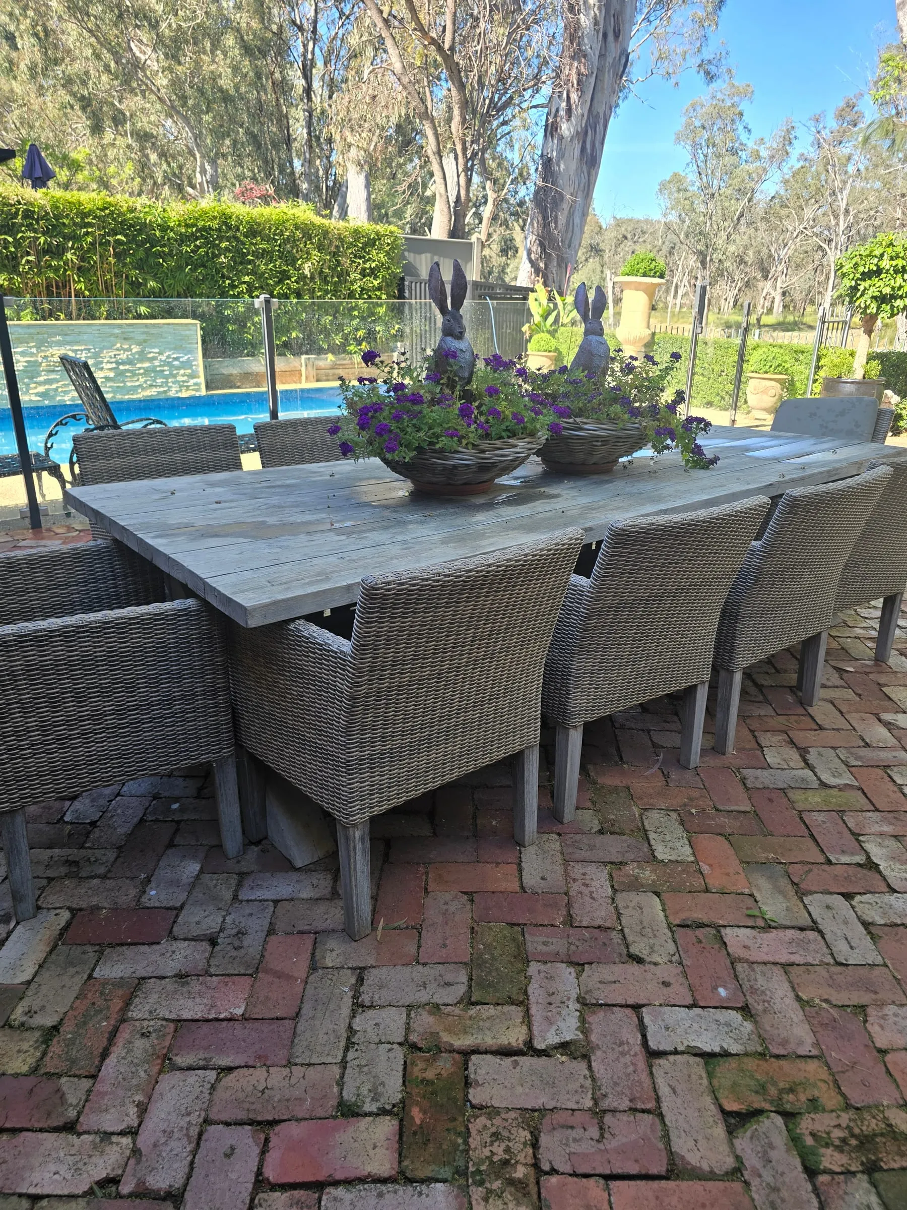 Long timber table and wicker chairs on the brick courtyard, set with purple flowers, the pool and river gums beyond